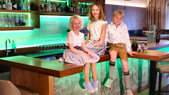 Drei lächelnde Kinder in traditioneller bayerischer Kleidung auf einer modernen Bartheke